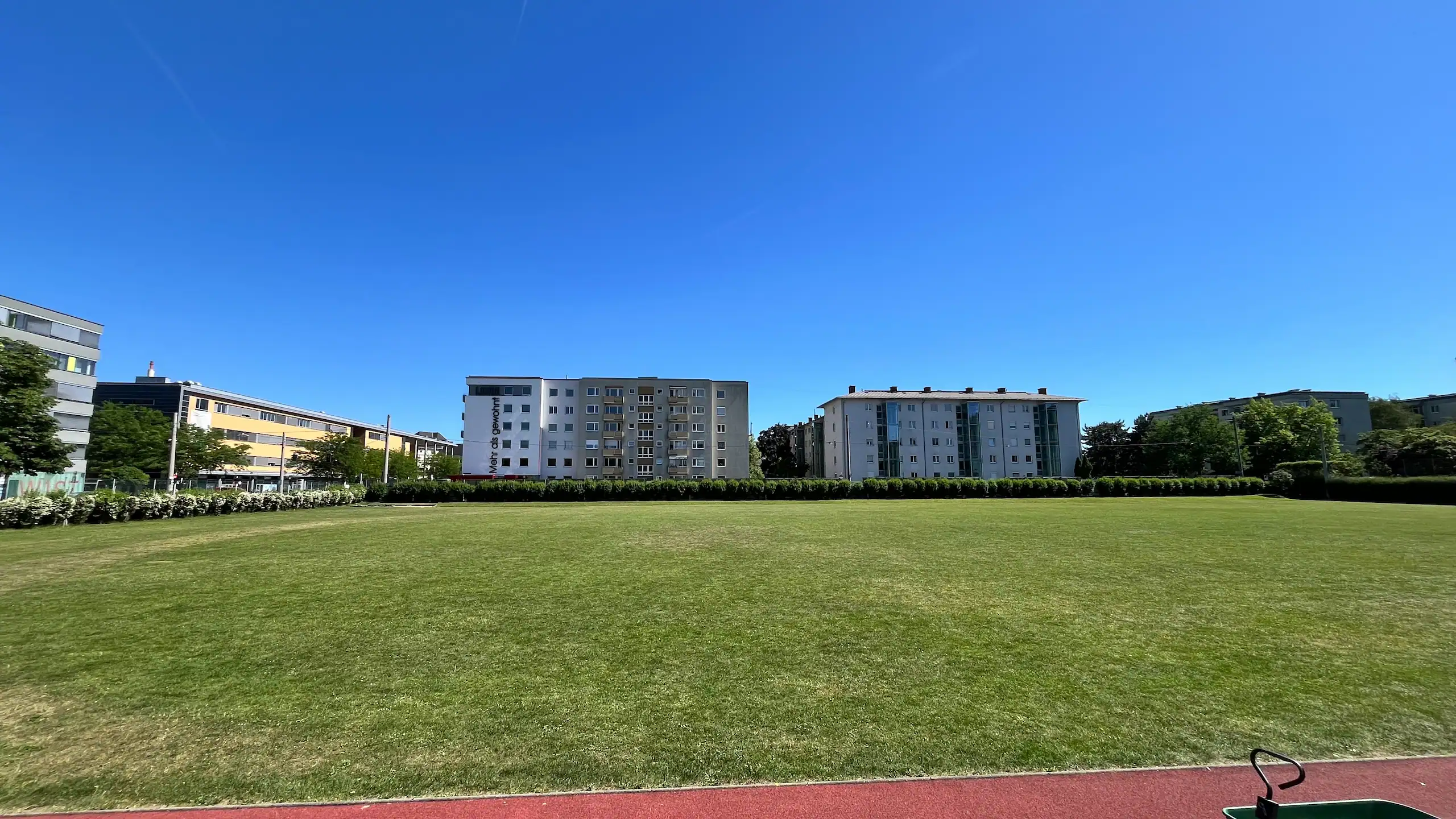 Panorama-Aufnahme der weitläufigen Sportanlage des ASKÖ Linz-Stamm an einem sonnigen Tag. Im unteren Vordergrund ist ein Streifen der roten Laufbahn zu sehen, gefolgt von einer großen, gepflegten Rasenfläche. Den Hintergrund bilden städtische Wohnblocks unter einem tiefblauen, wolkenlosen Himmel, die durch eine dichte grüne Hecke vom Sportplatz abgegrenzt sind.